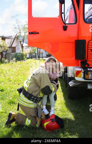 Firefighter in uniform with rescued little girl near fire truck outdoors. Save life Stock Photo