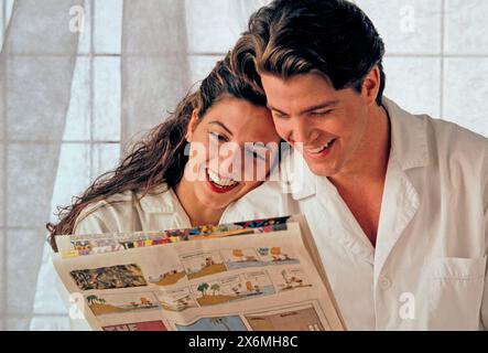 A man and a woman in pajamas are smiling as they read the Sunday comics together. They are both seated, engrossed in the content, with expressions of Stock Photo