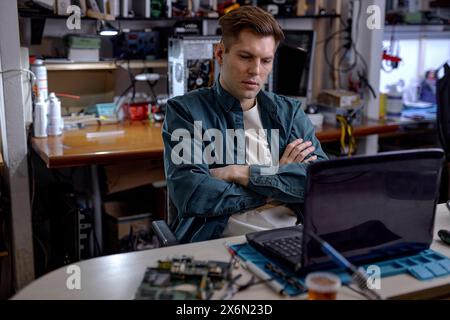 caucasian professional technician sit thinking how to repair fault laptop, sit behind table at workshop. Repairs and diagnostics breakdowns of laptop in professional service center. Stock Photo