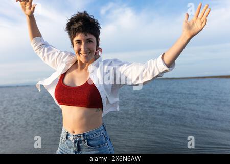 short-haired woman smiling on the beach, wearing white shirt, red top and denim, blue sky and calm sea. Stock Photo