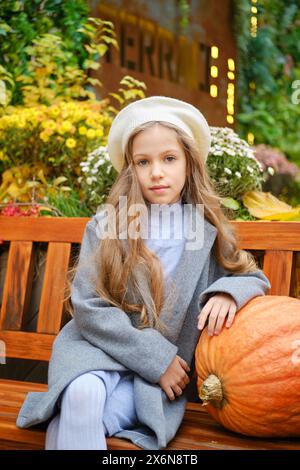 Cute little girl in a beret and woolen coat sits on bench leaning on pumpkin Stock Photo