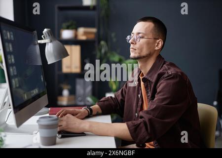 Minimal side view portrait of young Asian man using computer at workplace and typing against black wall Stock Photo