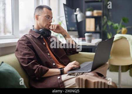 Side view portrait of adult Asian man wearing glasses and using laptop sitting on couch in office and working in IT copy space Stock Photo