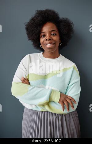 Vertical portrait of smiling African American woman with natural hair standing against grey wall dressed in casual clothes and looking at camera Stock Photo