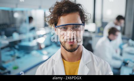 Medical Science Laboratory: Handsome Young Latin Scientist Wearing White Coat and Safety Glasses Smiles Looking at Camera. Young Bio Technology Research Specialist. Medium Closeup Portrait Shot Stock Photo