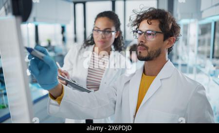 Medical Science Laboratory: Handsome Latin Male Scientist Writes Detailed Project Data Analysis on the Board, His Black Female Colleague Talks. Young Scientists Solving Problems. Stock Photo
