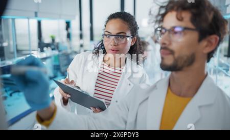 Medical Science Laboratory: Handsome Latin Male Scientist Writes Detailed Project Data Analysis on the Board, His Black Female Colleague Talks. Young Scientists Solving Problems. Stock Photo