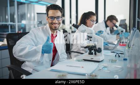 Medical Science Laboratory: Handsome Scientist is Using Microscope for Sample Analysis, Looking at Camera, Smiling Cheerfully, Shows Thumbs Up. Young Biotechnology Specialist, Using Advanced Equipment Stock Photo