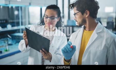 Medical Science Laboratory: Handsome Latin Male Scientist Doing Project Data Analysis, His Black Female Colleague Checks Information on Clipboard. Young Scientists Solving Problems Stock Photo
