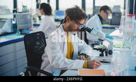 Medical Science Laboratory: Handsome Latin Scientist is Using Microscope for Sample Analysis and Writes Down the Result, Data. Young Biotechnology Specialist, Using Advanced Equipment. Stock Photo