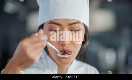 World Famous Restaurant: Portrait of Smiling Asian Female Chef Cooking Delicious, Steamy, Authentic Food, Tastes Her Dish With a Spoon in a Kitchen. Preparing affordable Haute Cuisine gourmet Stock Photo