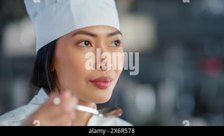 World Famous Restaurant: Portrait of Smiling Asian Female Chef Cooking Delicious, Steamy, Authentic Food, Tastes Her Dish With a Spoon in a Kitchen. Preparing affordable Haute Cuisine gourmet Stock Photo