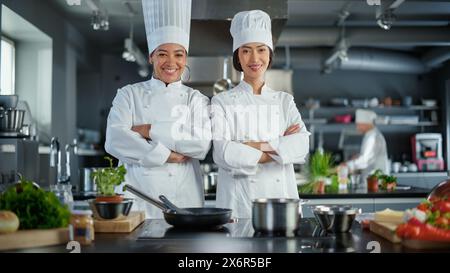 World Famous Restaurant: Portrait of Asian and Black Female Chefs Posing, Looking at Camera.Two Professionals Cooking Delicious and Authentic Food, Preparing Healthy Meals in Modern Kitchen. Wide Shot Stock Photo