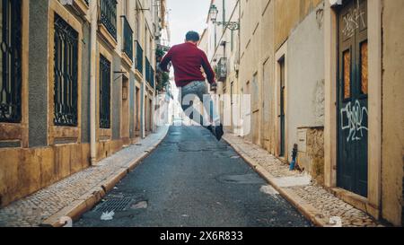 Cheerful and Happy Young Adult Man in Casual Clothes Actively Dancing while Walking on the Street of an Old Town in a City. Scene Shot in an Urban Quiet Narrow Town Street. Shot from Back. Stock Photo
