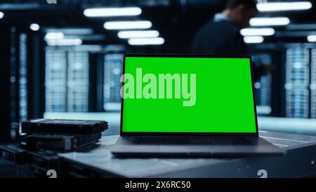 Laptop Computer with Green Screen Chroma Key Mock Up Display Stands on a Table. In the Background Data Center With Rows of Server Racks and IT Engineer Working. Advanced Equipment. Stock Photo