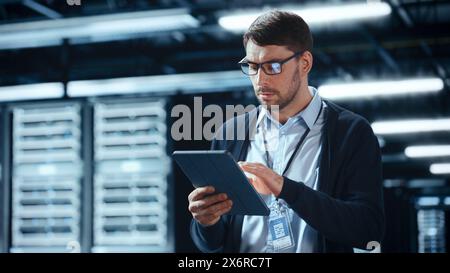 Male IT Specialist Walks Between Row of Operational Server Racks in Data Center. Engineer Uses Tablet Computer for Maintenance. Concept for Cloud Computing, Artificial Intelligence, Cybersecurity. Stock Photo