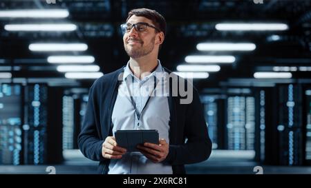 Male IT Specialist Walks Between Row of Operational Server Racks in Data Center. Engineer Uses Tablet Computer for Maintenance. Concept for Cloud Computing, Artificial Intelligence, Cybersecurity. Stock Photo