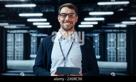 Portrait of a Bearded Handsome Caucasian IT Specialist in Glasses Standing with Tablet and Posing in the Middle of a Working Data Center Server Room with Server Computers Working on a Rack. Stock Photo