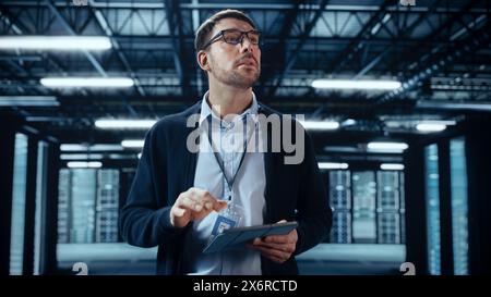 Male IT Specialist Walks Between Row of Operational Server Racks in Data Center. Engineer Uses Tablet Computer for Maintenance and Diagnostics in Cyber Security and Data Protection Facility. Stock Photo