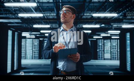 Male IT Specialist Walks Between Row of Operational Server Racks in Data Center. Engineer Uses Tablet Computer for Maintenance and Diagnostics in Cyber Security and Data Protection Facility. Stock Photo