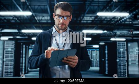 Male IT Specialist Walks Between Row of Operational Server Racks in Data Center. Engineer Uses Tablet Computer for Maintenance. Concept for Cloud Computing, Artificial Intelligence, Cybersecurity. Stock Photo