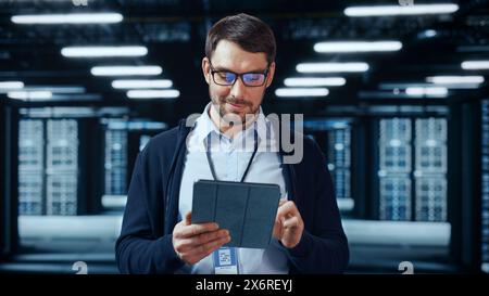 Portrait of a Bearded Handsome Caucasian IT Specialist in Glasses Standing with Tablet and Posing in the Middle of a Working Data Center Server Room with Server Computers Working on a Rack. Stock Photo