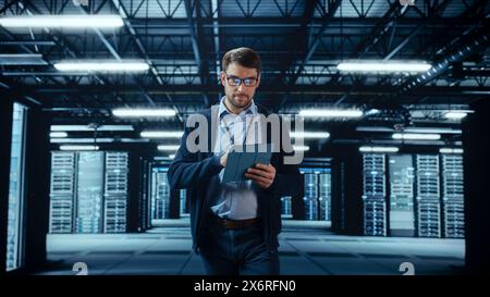Male IT Specialist Walks Between Row of Operational Server Racks in Data Center. Engineer Uses Tablet Computer for Maintenance. Concept for Cloud Computing, Artificial Intelligence, Cybersecurity. Stock Photo