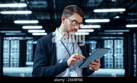 Portrait of a Bearded Handsome Caucasian IT Engineer in Glasses Standing with Tablet and Posing in the Middle of a Working Data Center Server Room with Server Computers Working on a Rack. Stock Photo