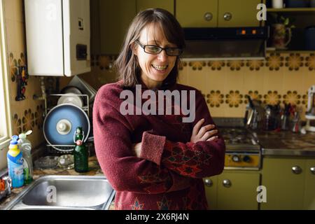 An Adult Woman in a red sweater is thinking looking down stands in front of a kitchen sink. She is calm, thinking and contemplating with her arms cros Stock Photo