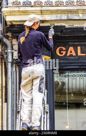 Young female decorator sanding shop front prior to repainting - Loches, Indre-et-Loire (37), France. Stock Photo