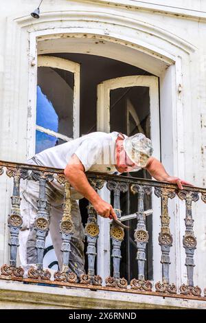 Workman using wire brush to scrape loose paint and rust from balcony  icast-iron railing - Loches, Indre-et-Loire (37), France. Stock Photo
