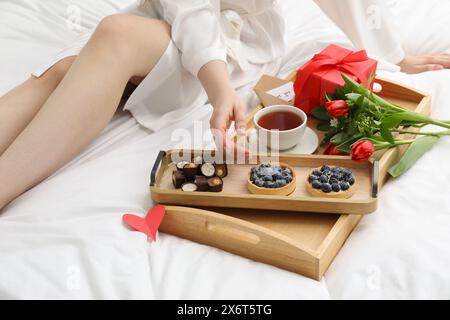 Tasty breakfast served in bed. Woman with desserts, tea, gift box and flowers at home, closeup Stock Photo