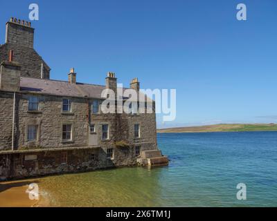 Old stone building on the waterfront with clear skies and calm sea in the background, calm sea with rocks and fields with historic houses, Lerwick Stock Photo