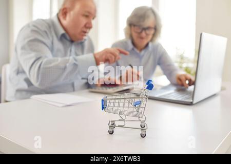 Close Up of a Toy Shopping Cart with an Old Couple Working on a Laptop at Home Stock Photo