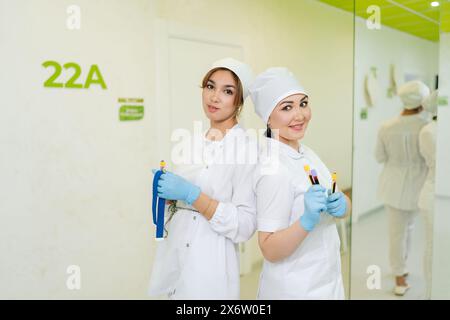 Two friendly medical workers holding test tubes with blood standing on hallway in laboratory. Charming nurses in medical uniform and blue gloves shows Stock Photo