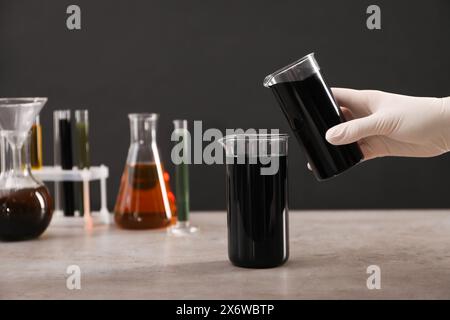 Woman pouring black crude oil into beaker at grey table against dark background, closeup Stock Photo