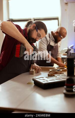 Woodworker next to BIPOC coworker using power drill to create holes for dowels in wooden board. Carpenter working with apprentice, sinking screws into wooden surfaces with electric tool Stock Photo