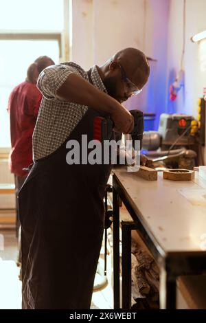 African american carpenter drills hole into timber block, doing furniture assembling in studio. BIPOC woodworking specialist inserting nail into piece of wood using electric power drill tool Stock Photo