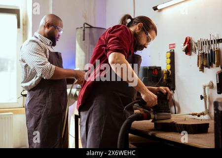 Carpenter collaborating with african american apprentice using orbital sander with fine sandpaper to achieve refined finish. Man and coworker using angle grinder on wood block for professional results Stock Photo