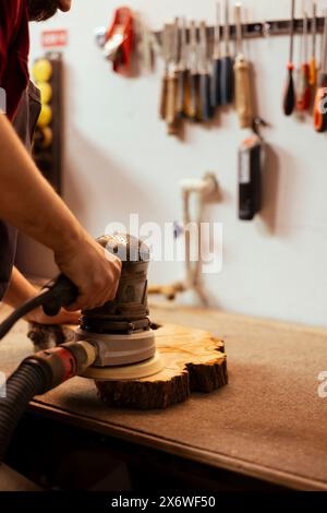 Cabinetmaker at workbench using orbital sander with fine sandpaper on lumber to achieve smooth finish. Woodworking specialist in assembly shop uses angle grinder on wood for professional results Stock Photo