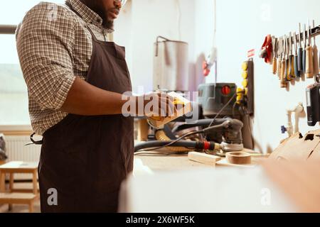 African american man in assembly shop using sandpaper for sanding wooden surface before painting it, ensuring adequate finish. BIPOC carpenter using abrasive sponge to repair damages on wood block Stock Photo