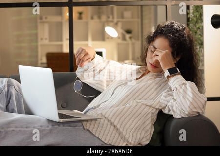 Tired female African-American programmer with laptop lying on sofa in office at night Stock Photo