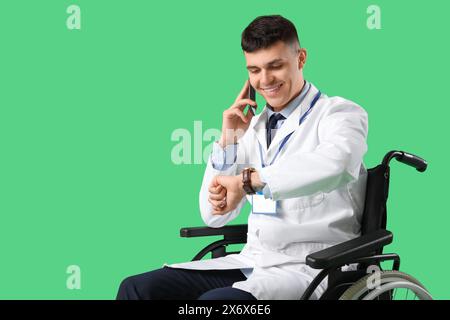 Young male doctor in wheelchair talking by phone and checking time on green background Stock Photo