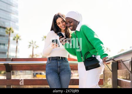 two young female friends smiling happy while having fun carefree looking at their mobile phones in a city park, concept of diversity and modern lifest Stock Photo