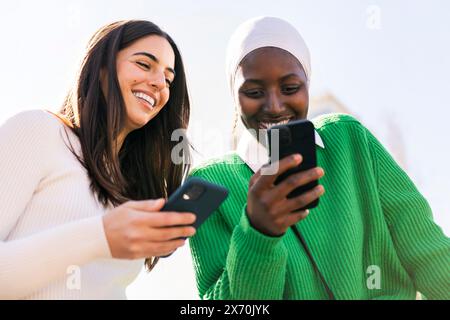 two young female friends smiling happy while having fun carefree looking at their mobile phones, concept of diversity and modern lifestyle Stock Photo