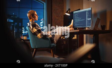 Young Woman Writing Code on Computer in Stylish Loft Apartment in the Evening. Creative Female Wearing Cozy Clothes, Resting Feet on Table and Using Wireless Keyboard. Urban City View from Big Window. Stock Photo