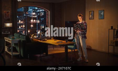 Young Beuatiful Women Working from Home, Standing And Holding Laptop Computer in Stylish Loft Apartment in the Evening. Creative Female Wearing Cozy Casual Outfit. Urban City View from Big Window. Stock Photo