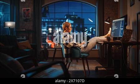 Young Woman Writing Code on Computer in Stylish Loft Apartment in the Evening. Creative Female Wearing Cozy Clothes, Resting Feet on Table and Using Wireless Keyboard. Urban City View from Big Window. Stock Photo