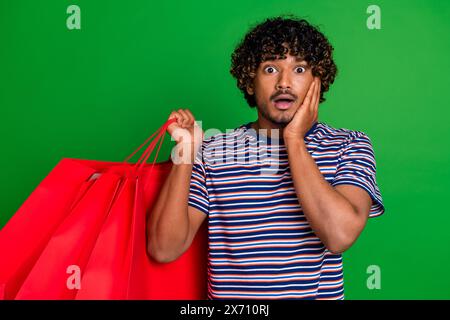 Portrait photo of young indian curly funny guy student touch cheek wearing striped t shirt stressed isolated on green color background Stock Photo