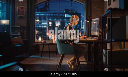 Young Woman Writing Code on Desktop Computer in Stylish Loft Apartment in the Evening. Creative Female Wearing Headphones, Working from Home on Software Development. Urban City View from Big Window. Stock Photo